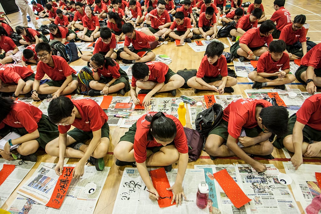 Students in red shirts sit on a gym floor practicing Chinese calligraphy on red paper spread over newspapers.